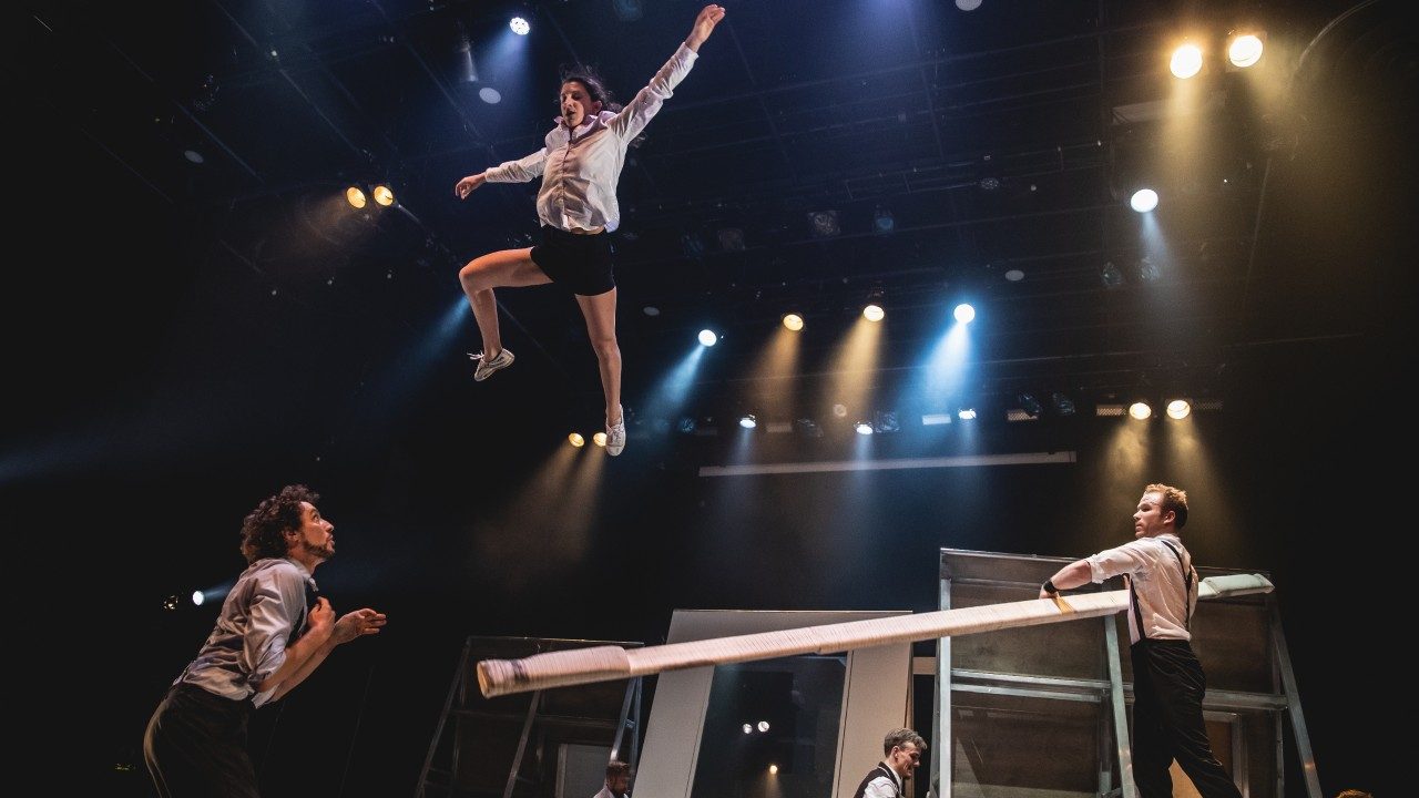  Members of Machine de Cirque perform on stage. On the right, a white man holds a long white wooden board. On the left, a woman in a light blue button down shirt and black shorts jumps through the air gracefully, and a man with brown curly hair waits on the ground below to catch her.