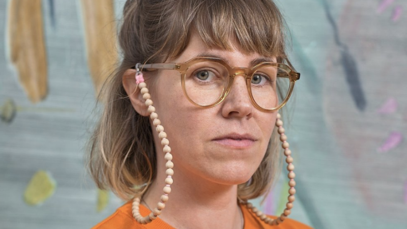 A portrait of artist Clare Grill, a young white woman with shoulder length sandy brown hair and brown round framed glasses. She wears an orange shirt and uses a beaded glasses chain. She poses in front of one of her works.