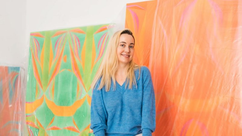 A portrait of artist Margaux Ogden, a white woman with long blonde hair. She wears a cornflower blue sweater and sits in front of some of her works in her studio.