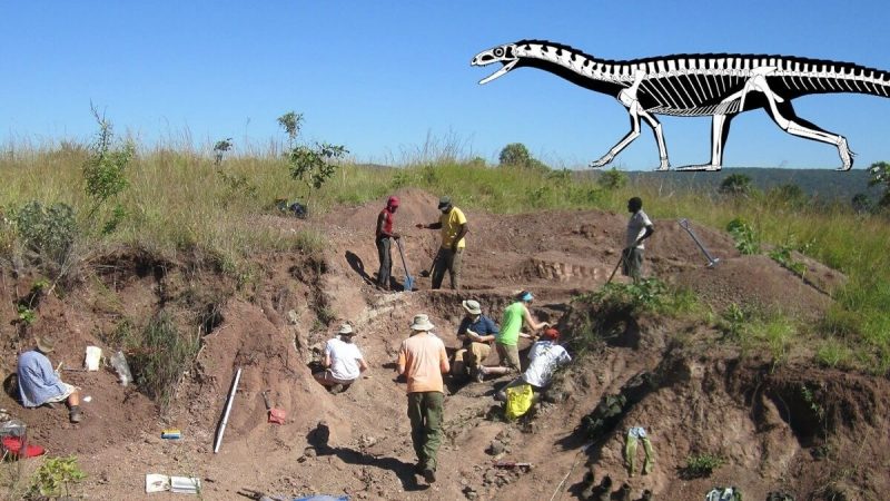  In the foreground, archaeologists work at a dig site on a sunny day. In the background is rolling mountains and the image of an animated dinosaur skeleton walking on the top of the mountain ridge.