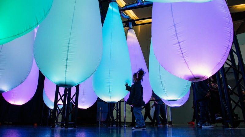  A young girl gently touches a light blue, teardrop-shaped balloon in an exhibition. She is surrounded by other blue, green, and purple balloons of the same shape and size.
