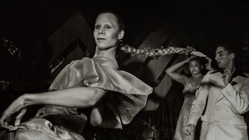  Larry Fink's "Studio 54, New York, NY, May 1977;" archival pigment print; 22 x 17 inches; courtesy of the Virginia Tech Art Collection and donors Scott and Emily Freund and Michael Fay. In this black and white photo, a white woman twirls on the dance floor. She is wearing an off-the-shoulder dress with ruffles at the arms and heavy make up. Her long blonde hair is pulled back into a tight braided ponytail, which is flying out in midair behind her. In the background are two other dancers, a Black woman in a spaghetti strap dress and a white man in a white power suit.