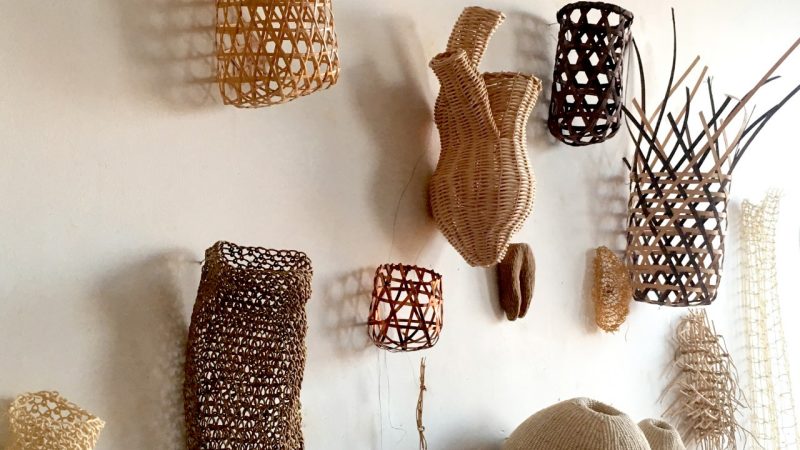  Miscellaneous handmade baskets hang on a pale beige wall. The piece is "ephemera," 2015-2021, by Ann Coddington.