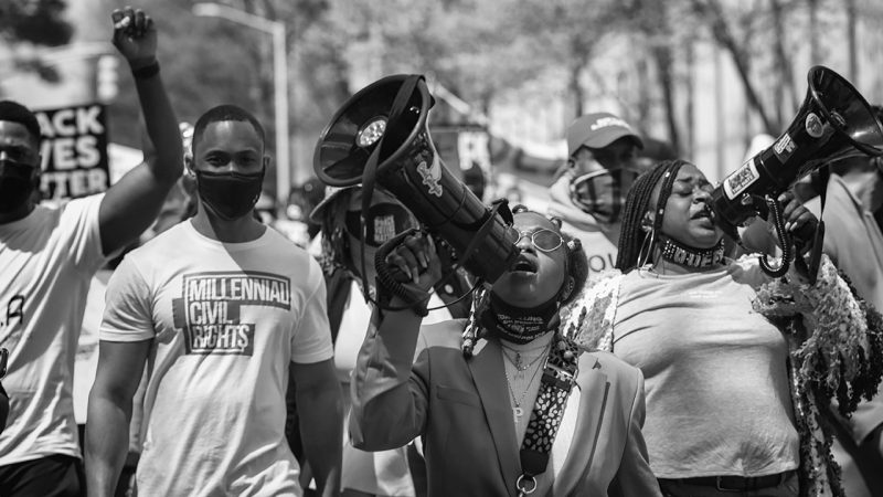  Sheila Pree Bright's "The People's Uprising, Jim Crow 2.0 Voter Suppression Rally, Atlanta, GA;" digital print; 36 x 24 inches; courtesy of the artist. Pictured are several young Black men and women speaking into megaphones, wearing masks, and protesting in the streets.