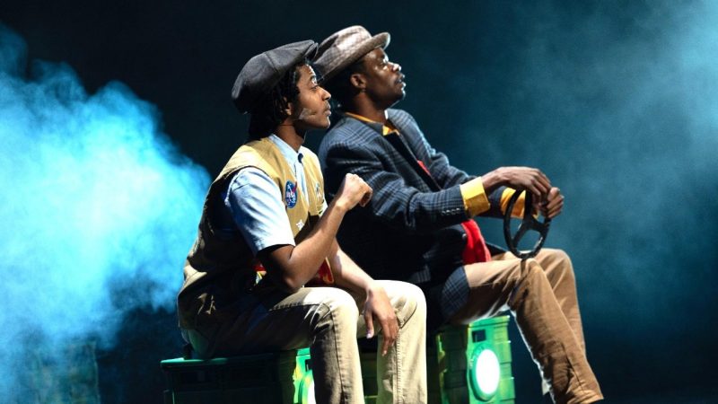 Two actors from "Hero: The Boy from Troy" perform on stage, as if sitting together in a car. Theatrical haze floats behind them.