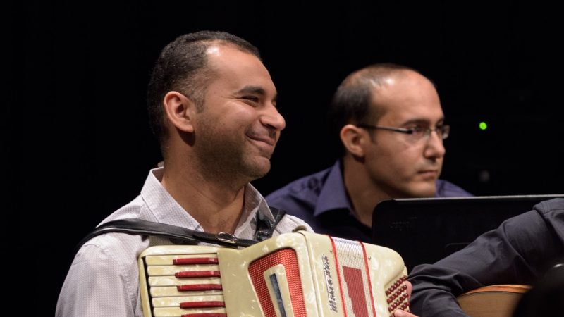 Mohammed Seyam, a Middle Eastern man, smiles while on stage with the Itraab Arabic Music Ensemble, an accordion in his lap.