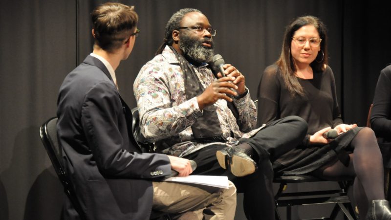  Three people sitting in a row in the Cube for a lecture series. The middle gentleman, Awadagin Pratt, wears a print collared shift with black scarf and holds a microphone while speaking.