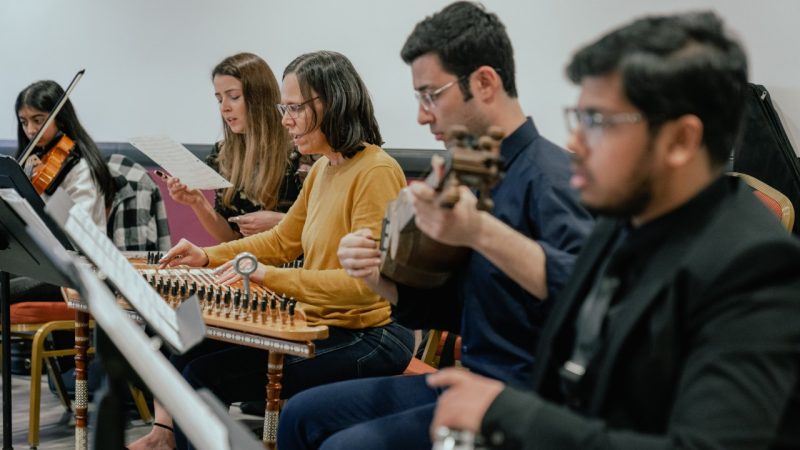 The Itraab Arabic Music Ensemble performs at the Moss Arts Center. In the foreground, from left, is a brown woman with dark brown hair pulled up into a chignon, red lipstick, and a black sweater and skirt playing a drum in her lap; a brown man playing a tambourine; and a brown man with a bald head and full dark brown beard playing a drum in his lap.