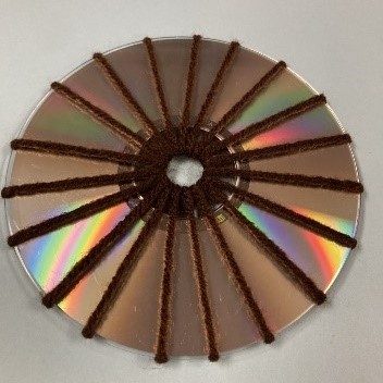 Image corresponding with step two of the CD weaving instructions. The shiny side of a CD is visible with an odd number of spokes wrapped around the CD in brown yarn.