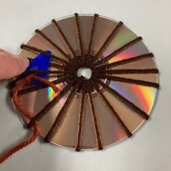 Image corresponding with step five of the CD weaving project. A white woman's hand threads a lighter brown piece of yarn through darker brown yarn spokes using a blue plastic needle. The yarn is wrapped around a CD, shiny side up.