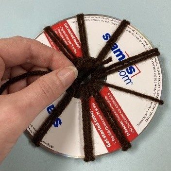 Image corresponding with step three of the CD weaving project. A white woman's hand ties a knot in brown yarn wrapped around an old CD.