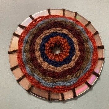 Image corresponding with step seven of the CD weaving project. The finished product, with yarn of lighter brown, dark blue, beige, dark brown, taupe, grey, and red, spiraling out from the center of an old CD. About a half an inch of the shiny side is visible along the edges.