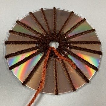 Image corresponding with step four of the CD weaving project. An odd number of dark brown yarn spokes looped through the center hole of a CD, shiny side facing out. A lighter brown piece of yarn is tied to the center and trails away from the CD.