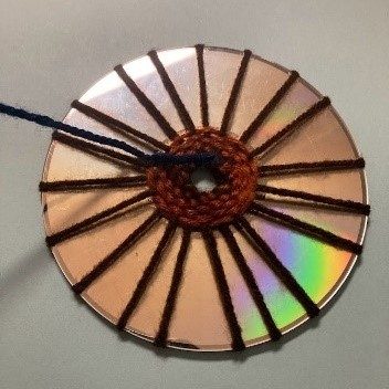 Image corresponding with step six of the CD weaving project. Lighter brown yarn is woven through darker brown yarn spokes, wrapped around an old CD, shiny side up. A new color of yarn, black or dark blue, has been tied to the end of the last bit of color, ready to continue weaving.