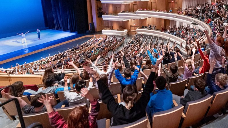  A crowd full of area PK-12 students participate during a school-day performance, raising their arms overhead in V shapes at the direction of a dancer on stage.