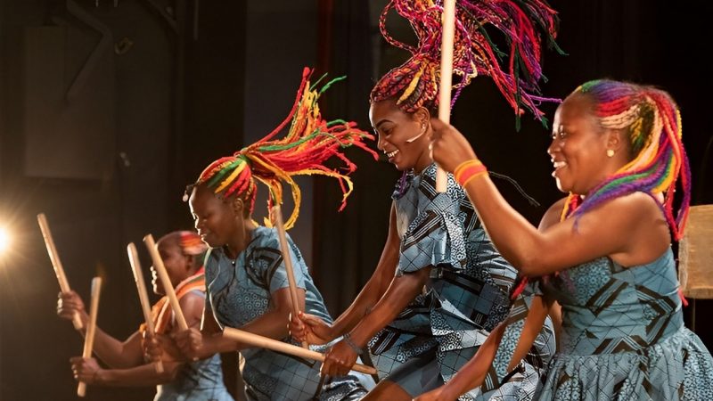  Women drummers from Rwanda perform onstage. They are Black women with brightly colored long braids, some of which are flying through the air with their motion. They wear blue and brown patterned dresses in slightly different styles.