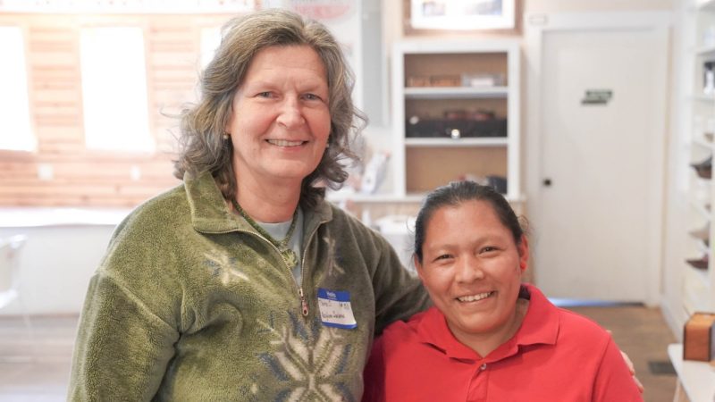  Tara Orlando, a white woman with shoulder length greying brown hair wearing a moss green fleece jacket, stands with a refugee she helps through her work. The refugee is a brown skinned woman with dark brown hair pulled back into a ponytail, and she wears a red polo.