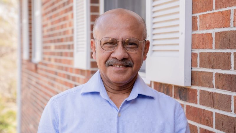  Jacob George, a brown man with a bald head, salt and pepper moustache, and round wireframed glasses, wears a blue button down shirt and stands in front of his home, a brick house with white shudders.