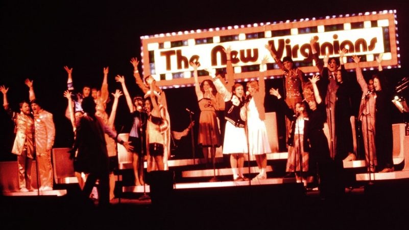  Members of the New Virginians perform on stage in this color image from 1981. The group wears an assortment of costumes and sings into microphones in pairs, one arm raised above their heads. Behind them is a large light-up New Virginians sign.
