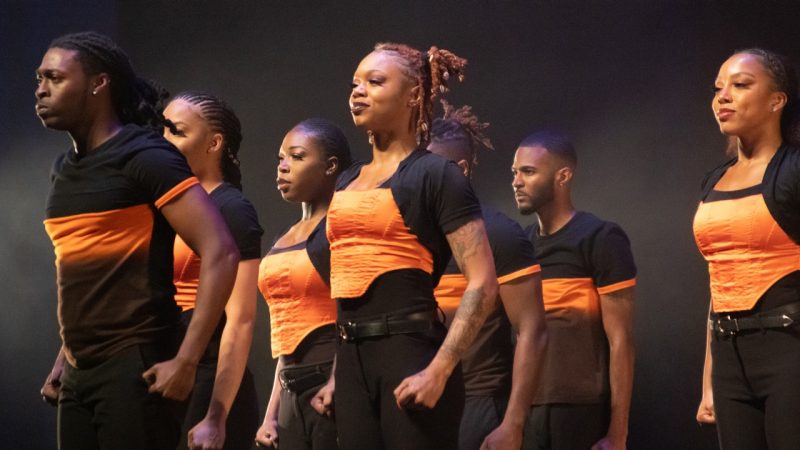 Members of Step Afrika! in orange and black costumes perform on stage.