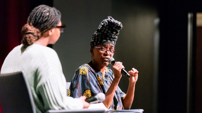 A Black woman wearing a patterned black head wrap, glasses, and a colorful patterned dress speaks into a microphone and looks towards a young Black woman in a mint green sweater wearing glasses, her braids pulled back into a low bun.