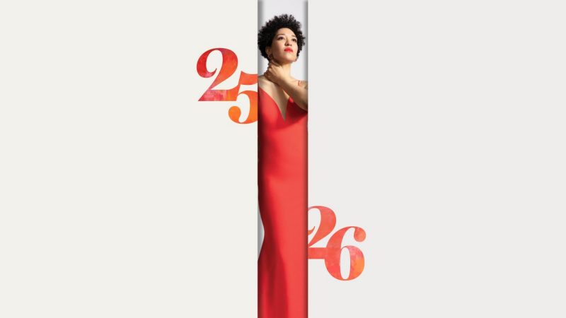2526season-performances Soprano Julia Bullock, a Black woman in a long red dress, stands in between two off-white panels, each with a red- and orange-toned 25 or 26.
