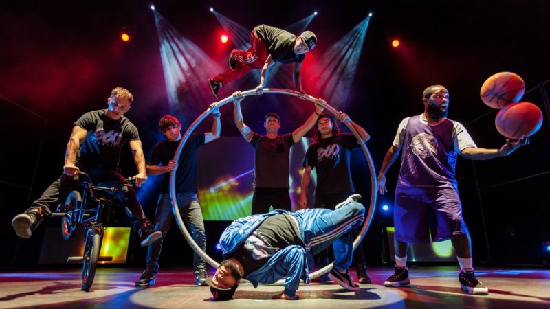 360-allstars_matt-loncar1 The cast of 360 ALLSTARS gathers on stage. From left, one rides a BMX bike, three hold up a large German wheel while a fourth man balances on top of it, a man hits a breakdance pose, and a man balances one basketball on top of another.