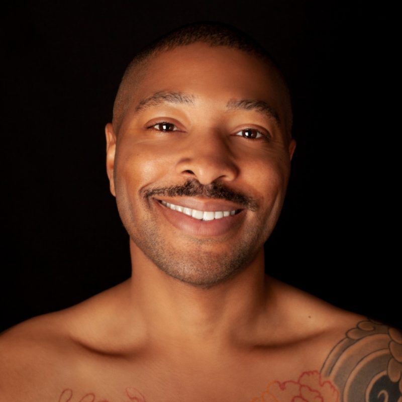 Company artistic director Kyle Abraham, of A.I.M by Kyle Abraham, smiles towards the camera against a black background in this headshot. Abraham is a young Black man with short natural hair, a moustache, and tattoos on his chest and left shoulder.