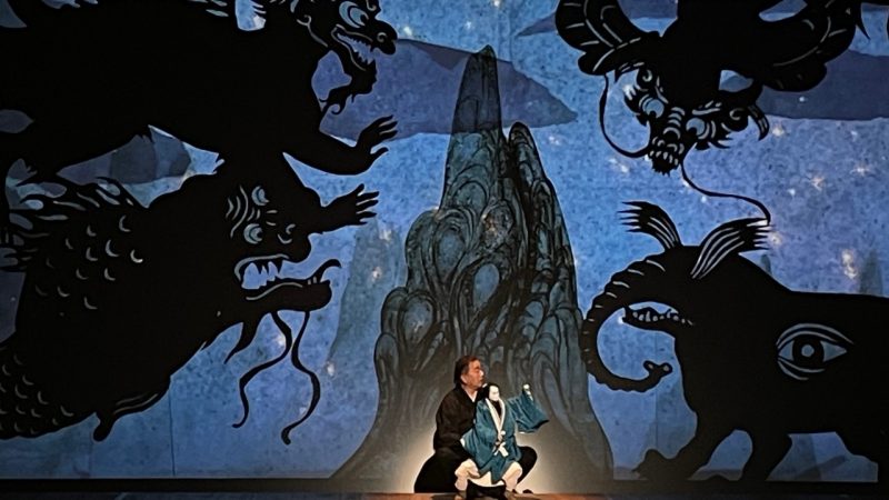  A Japanese man in black operates a large puppet, while illustrated monsters swirl on a projection behind them.