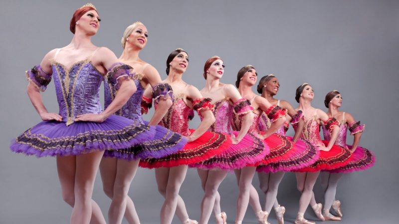  Eight of the dancers from Les Ballets Trockadero de Monte Carlo, all men dressed in beautiful, detailed drag. They wear tutus in shades of pink, purple, and red, and stand in a line, all in the same position with hands on their hips and looking towards the top right edge of the frame.