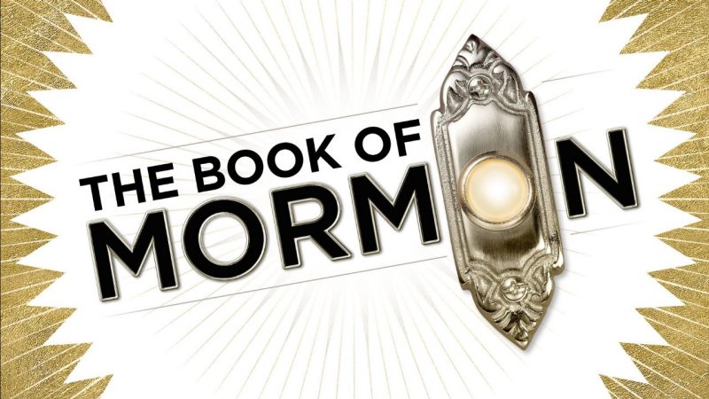 Two white men in white short sleeved button downs and black pants wave and lean into the frame. Between them, orange text on a white background reads "'The best musical of this century.' The New York Times"  The Book of Mormon logo, with the second O in Mormon replaced by a doorbell, is below with a faint starburst pattern surrounding it.