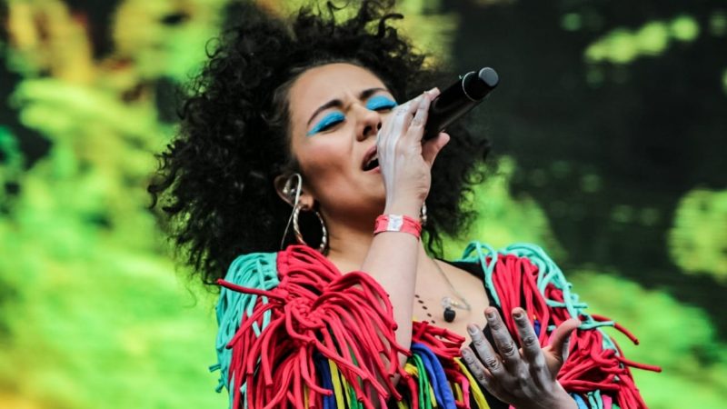  The lead singer for Chontadelia sings into a microphone with her eyes closed. She has natural curly brown hair and wears a turquoise and coral colored shirt with tassels, large gold hoop earrings, and electric blue eyeshadow. Her hands are painted a light grey
