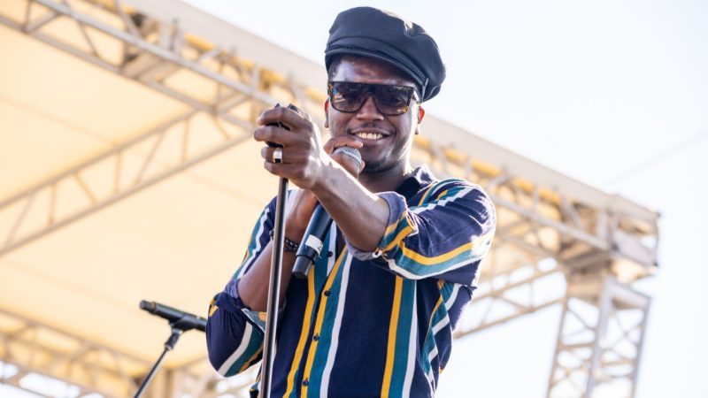  Musician Cimafunk, a Black man with a black hat, sunglasses, and blue and yellow vertically striped button down shirt, holds a microphone up to his mouth in one hand, the other hand resting on the top of a microphone stand. He's onstage of an outdoor venue during the day and is smiling towards the camera.