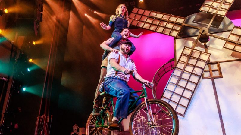  People ride three tall on a green bicycle on stage. Behind them is a large windmill, and a large pink backdrop.