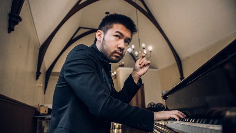  Conrad Tao, an Asian man with dark hair and a closely trimmed beard, sits at a piano in a dark blazer