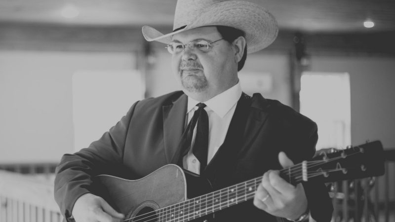  Junior Sisk of the Junior Sisk Band, a white man in a tan cowboy hat and black suit, holding an acoustic guitar. He has glasses and dark hair in this black and white image.