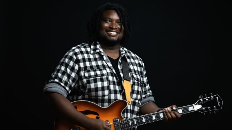  Justin Golden, a Black man with medium length dreads, smiles towards the camera, wearing a black and white buffalo check button down shirt, black T-shirt and holding a semi-hollow body guitar in front of a black background.