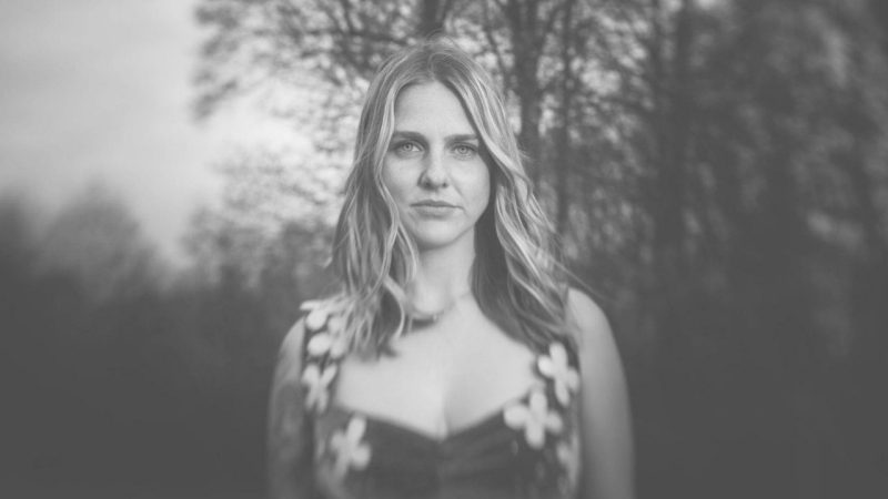  Dori Freeman, a white woman with medium length blonde hair, wears a dress with flower cutouts and stands in front of the edge of the woods in this black and white photo.