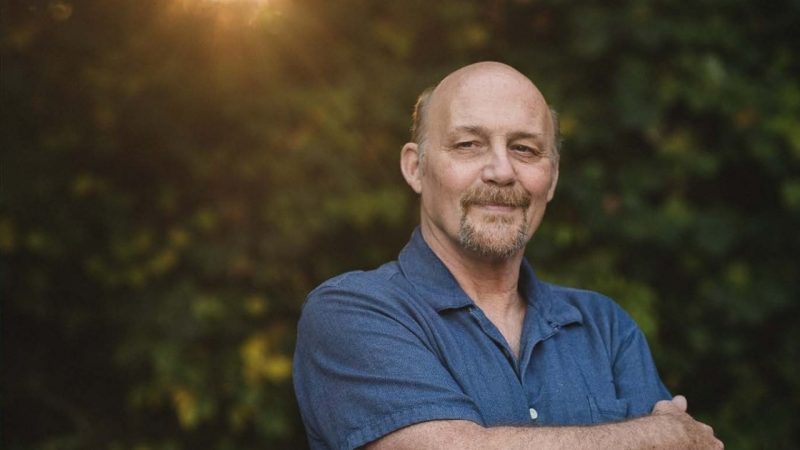  Eugene Wolf, narrator for the Crooked Road's Music and Dance Spectacular, is a white man with a balding head and light hair, a light brown goatee, and blue button down short sleeved shirt, standing in front of a patch of green trees, the Golden Hour sunlight streaming down behind him.