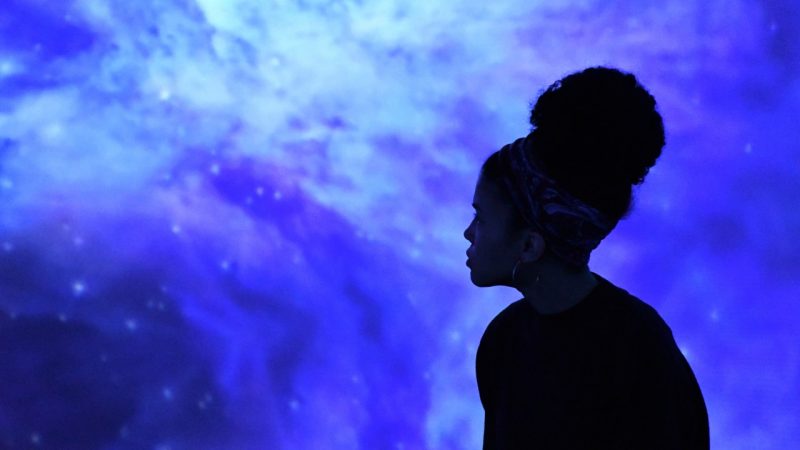 A woman listens to and experiences "Sound of Space" in the Cube at the Moss Arts Center. A black woman with a black shirt, natural hair pulled up into a bun on her head, and hoop earrings sits in profile against a swirling cosmic purple projection. 