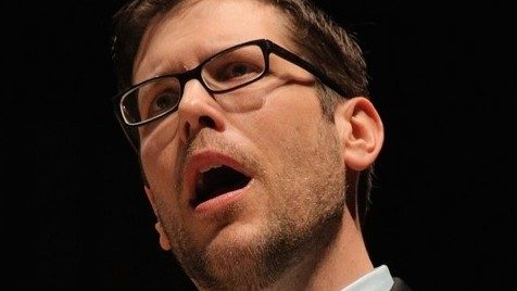 Tenor and Virginia Tech faculty member Brian Thorsett, a white man with short brown hair and black rectangular framed glasses, performs on stage in this close up.