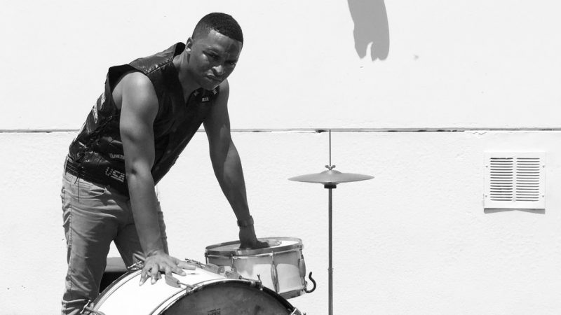 Drummer Deantoni Parks, a Black man with short naturla hair, wears a sleeveless black leather vest and jeans and leans on a drum kit. He poses outside in front of a white exterior wall.