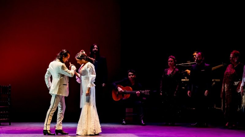  Flamenco dancer Farruquito and a female dancer wear all white and clasp hands on stage during a performance. Behind them, musicians and singers perform.