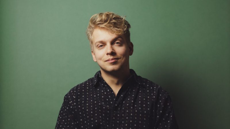 Countertenor Hugh Cutting, a young white man with wavy blonde hair, wearing a black patterned button down shirt in front of a green background.