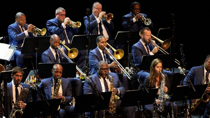  A promo photo of the Jazz at Lincoln Center Orchestra with Wynton Marsalis, near center. The group consists of 15 men in navy blue suits with brown shoes. They all stand or sit on stools in a row and hold their instruments in front of a white background.