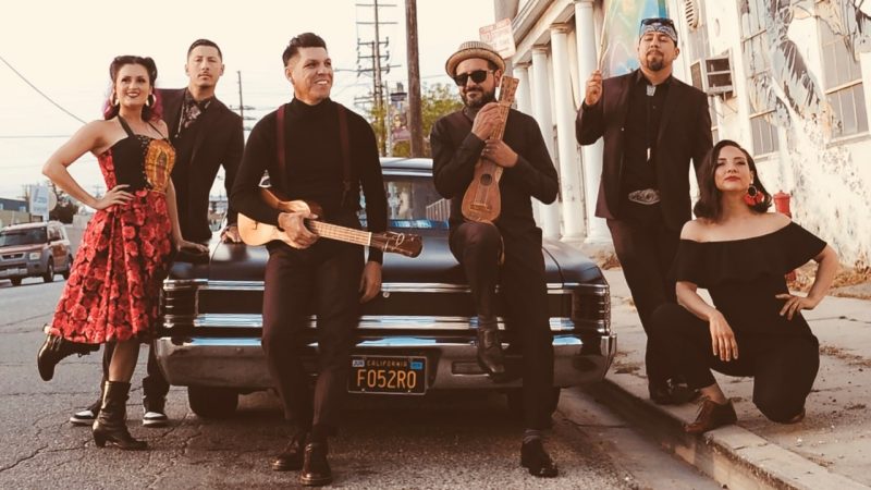  Members of East Los Angeles Chicano indie-folk band Las Cafeteras pose around a restored matte black El Camino. Some members hold Mexican ukulele-sized guitars and drum sticks. The four brown men wear all black and the two brown women wear black with red accents.