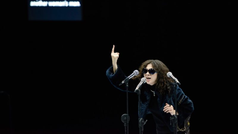  An actress, an Asian woman with long red-brown hair, wears sunglasses and a fur coat and speaks into three microphones in stands, her right hand pointing towards the ceiling.