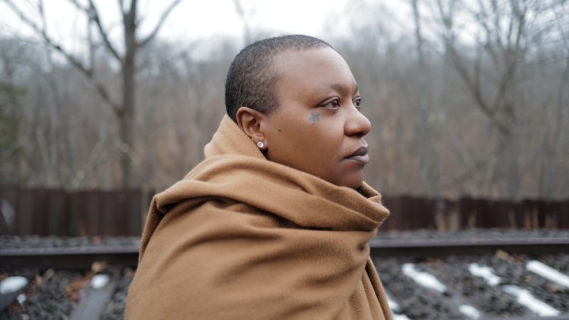  Musician Meshell Ndegeocello, a Black woman with shortly cropped natural hair and a star tatttooed near her right eye, stands outside near railroad tracks in the late fall or winter, wrapped in a camel-colored coat or large scarf. She looks towards the right of the frame, and some of the wooden tithes are covered in melting snow behind her.