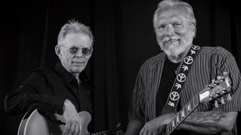  The members of Hot Tuna, two older white men with grey and white hair, smile towards the camera, each with a guitar hanging by its strap from their necks in this black and white image.