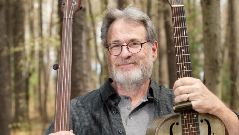 A middle aged white man with grey hair and beard wearing oval black framed glasses holds two acoustic instruments by their necks. He wears a grey button down over a grey shirt and is pictured in front of a forest.